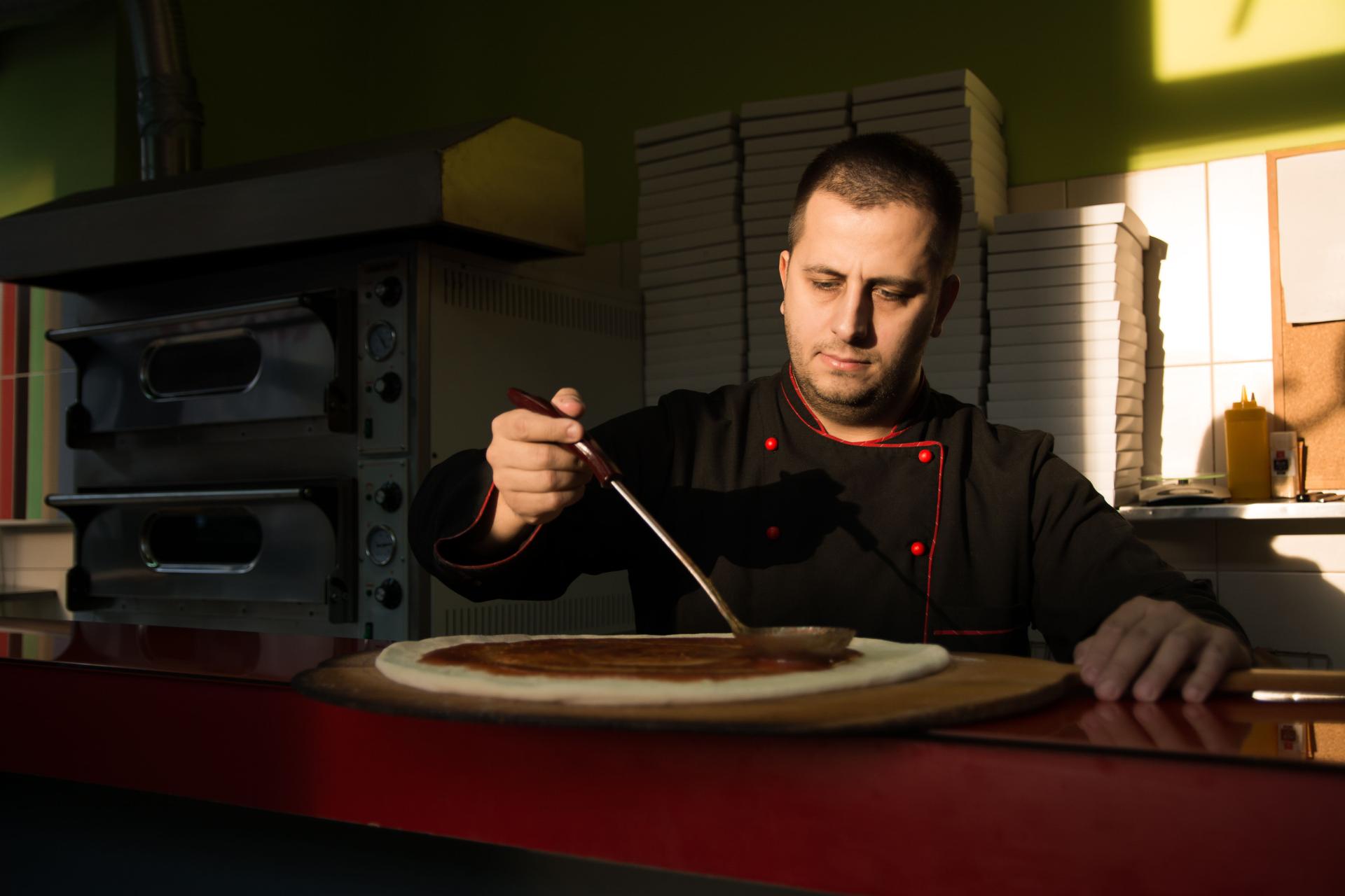 Pizza chef putting fresh tomato sauce on pizza base - Image